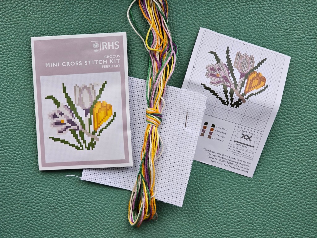 Contents of an assorted mini cross stitch kit. The small envelope includes thread in assorted colours, a small piece of canvas, a needle, and a leaflet with the pattern