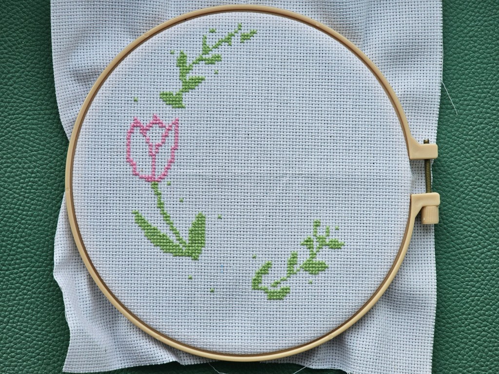 An embroidery hoop with mounted aida. A silhouette of a few flowers is sewn, but the design is very much in an early stage.