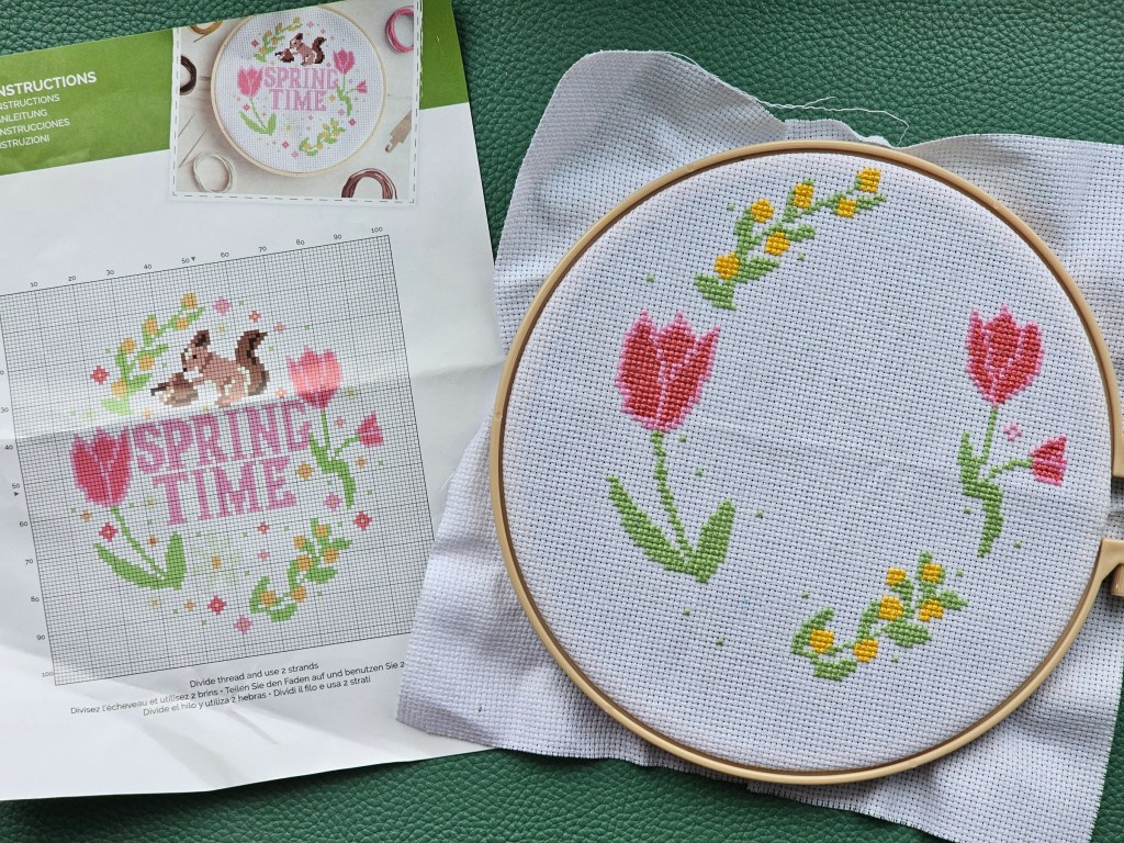 An embroidery hoop with mounted aida. A couple of pink tulips and some yellow flowers are sewn, but the centre of the design is empty.