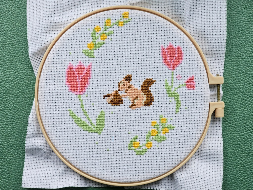 An embroidery hoop with mounted aida. A couple of pink tulips and some yellow flowers are sewn, and there's a squirrel in the centre.