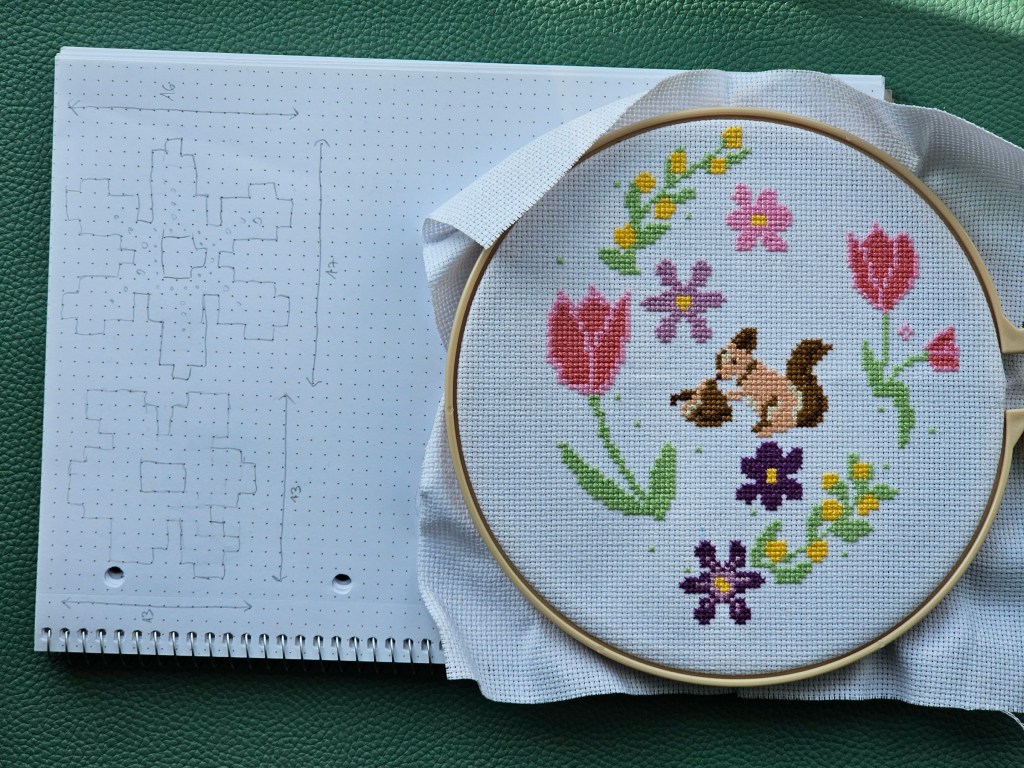 A notepad with two flower sketches, next to a cross stitch project with flowers and a squirrel.