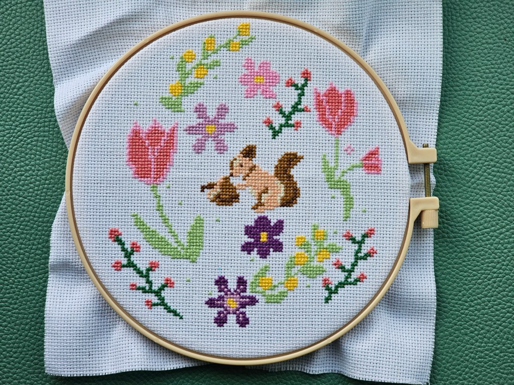 A nearly completed cross stitch design featuring a squirrel amongst colourful flowers.