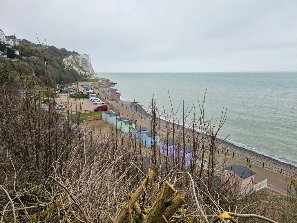 A row of colourful beach huts along the beach, seen from the top of a cliff.