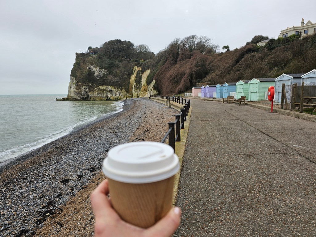 A hand holding a takeaway cup of coffee. Behind, tall white cliffs and a row of colourful beach huts can be seen.