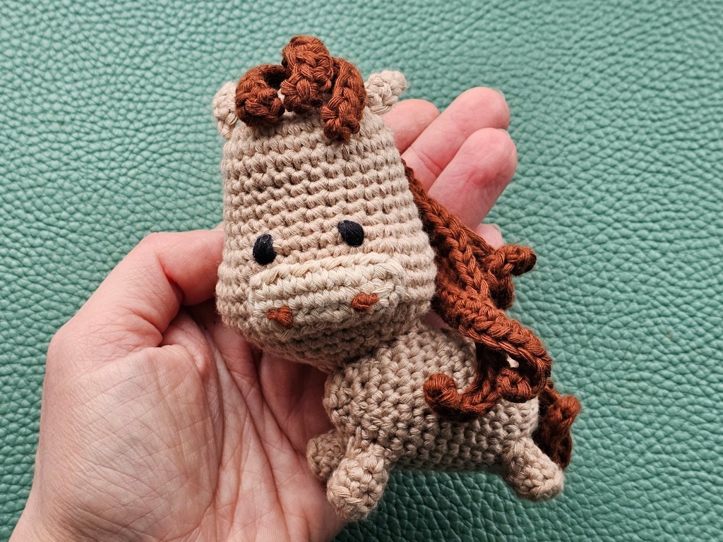 Completed small light brown crocheted pony toy, held on an open palm