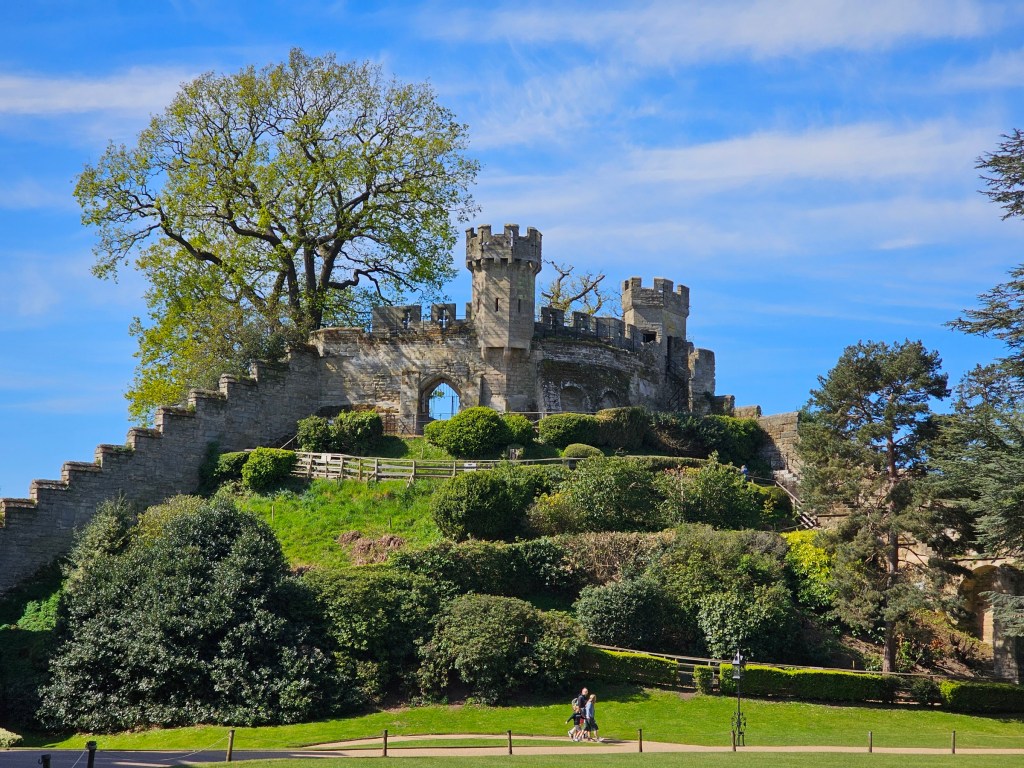 The mound of Warwick Castle. There's a small tower and a castle wall going up a small hill.