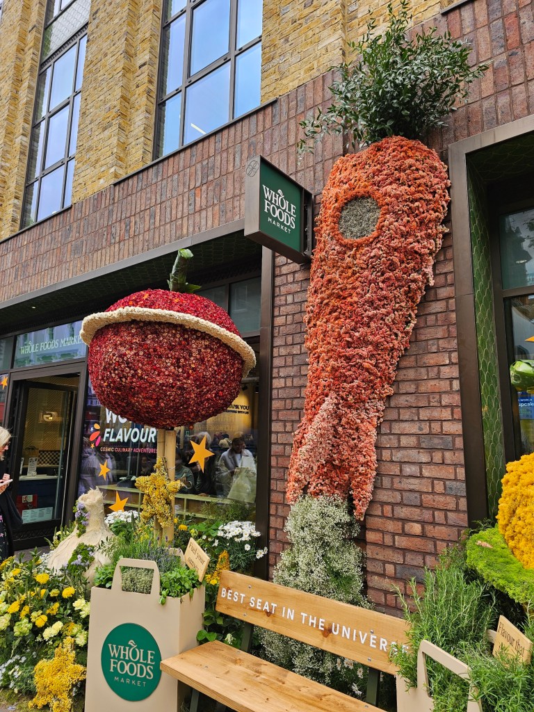 Statues of a planet-apple and spaceship-carrot made out of flowers. Photo taken during Chelsea in Bloom event in London in 2026