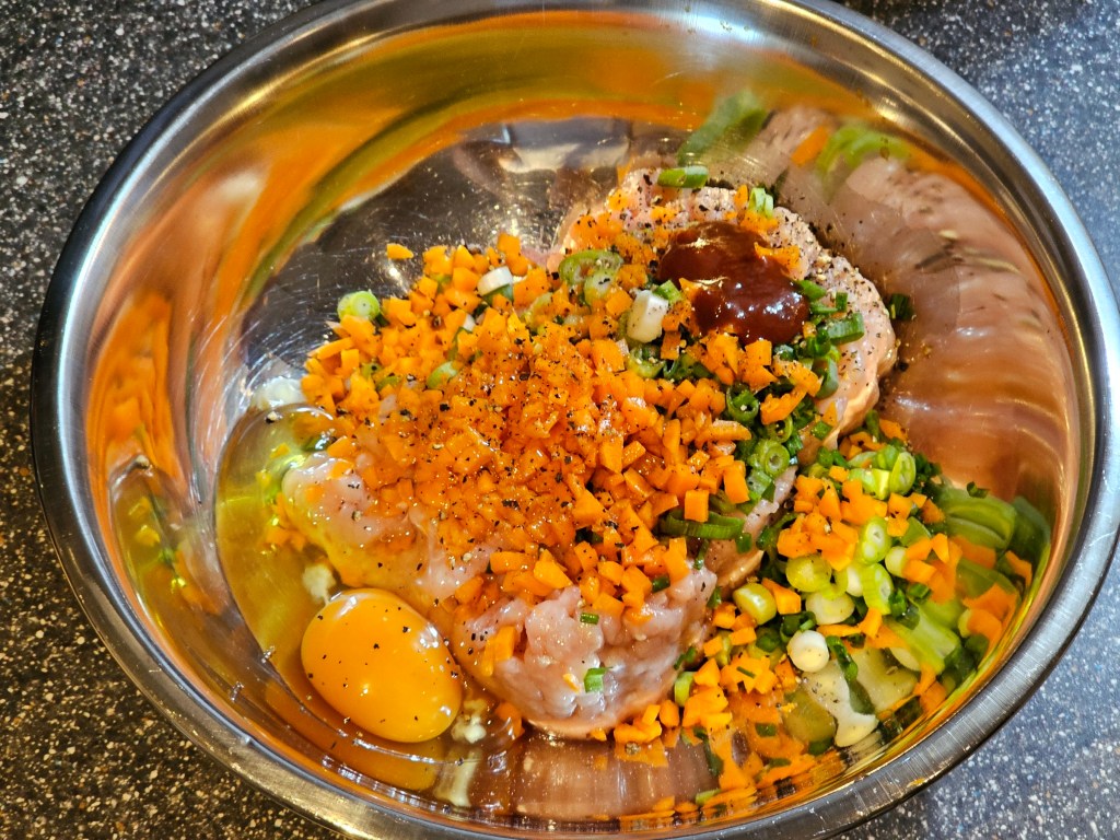 A metal bowl with ground chicken, an egg, diced carrot, chopped spring onions and hot sauce