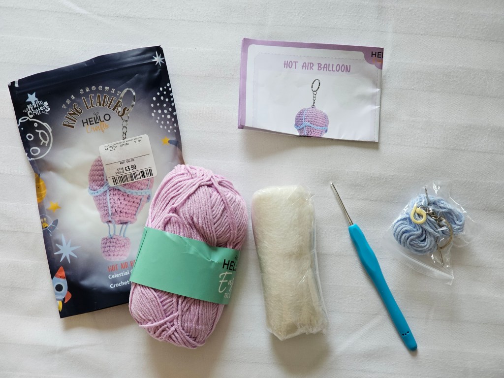 Contents of a DIY set for a crocheted hot air balloon. It includes pink and blue yarn, a crochet hook, a needle, a stitch marker, a keychain, toy stuffing and instructions