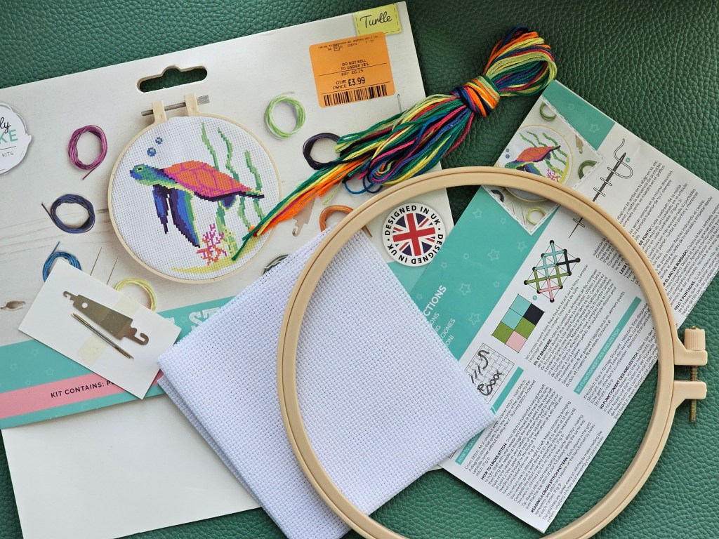 Contents of cross stitch set. It includes a plastic embroidery hoop, a couple of needles, a threader, a blank canvas, embroidery thread in several colours and a leaflet with instructions.