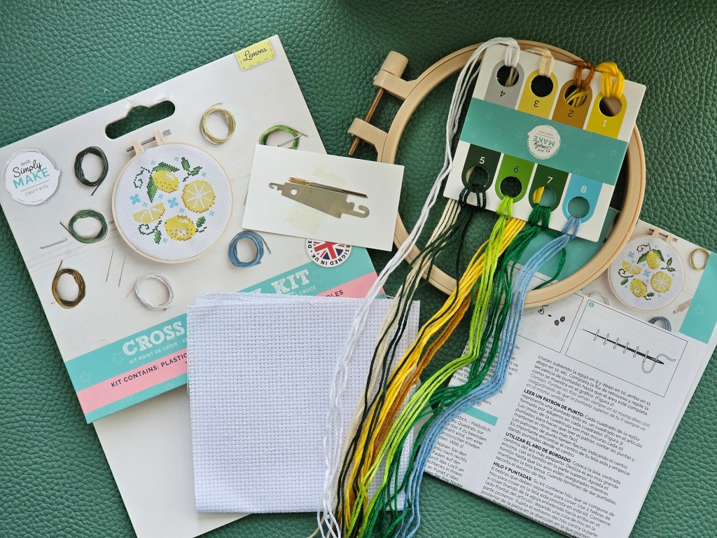 Contents of a mini cross stitch set with a lemon design. It includes a blank aida, embroidery thread in assorted colours, a needle, a threader, a hoop, and instructions