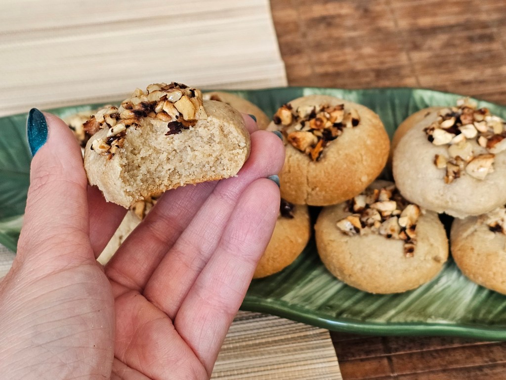 Baked tahini almond cookies topped with honey roasted nuts, on a leaf-shaped plate. A half eaten cookie is held in a hand.