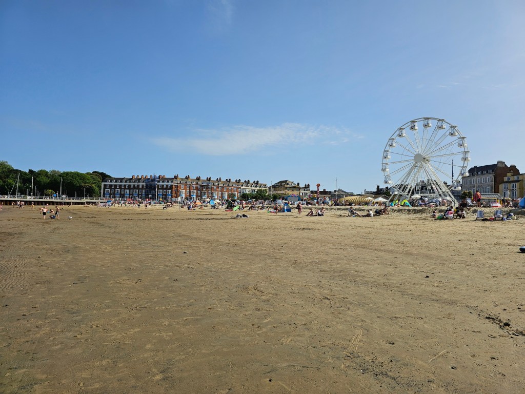Sandy beach in Weymouth, England. There's a lot of people sunbathing and a Ferris wheel