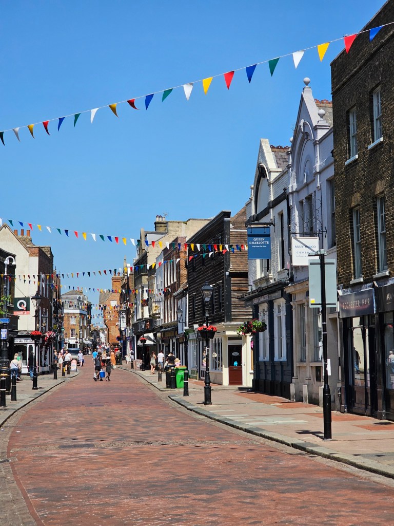 Rochester high street on a sunny day - there are old houses, each looking different than the others, and bunting hanging across the street