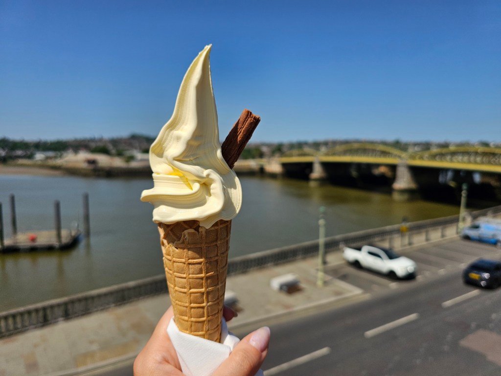 A soft serve ice cream cone with a piece of chocolate, held in a hand. A blurred bridge over a river in the background.