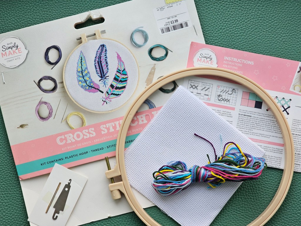 Contents of a cross stitch set: a plastic embroidery hoop, threads in assorted colours, a blank canvas, needles and a leaflet with the pattern.