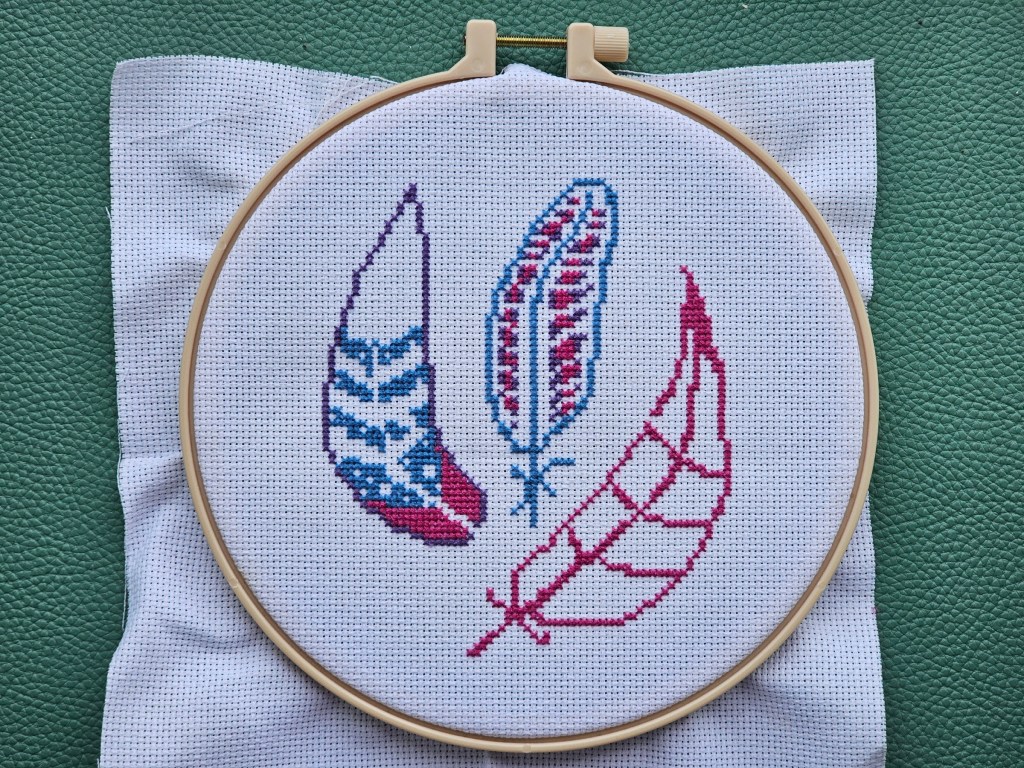 Partially finished cross stitch project featuring three pink, blue and purple feathers.