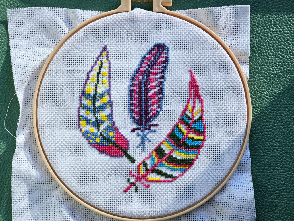Nearly finished cross stitch project featuring three pink, blue and purple feathers with yellow highlights.