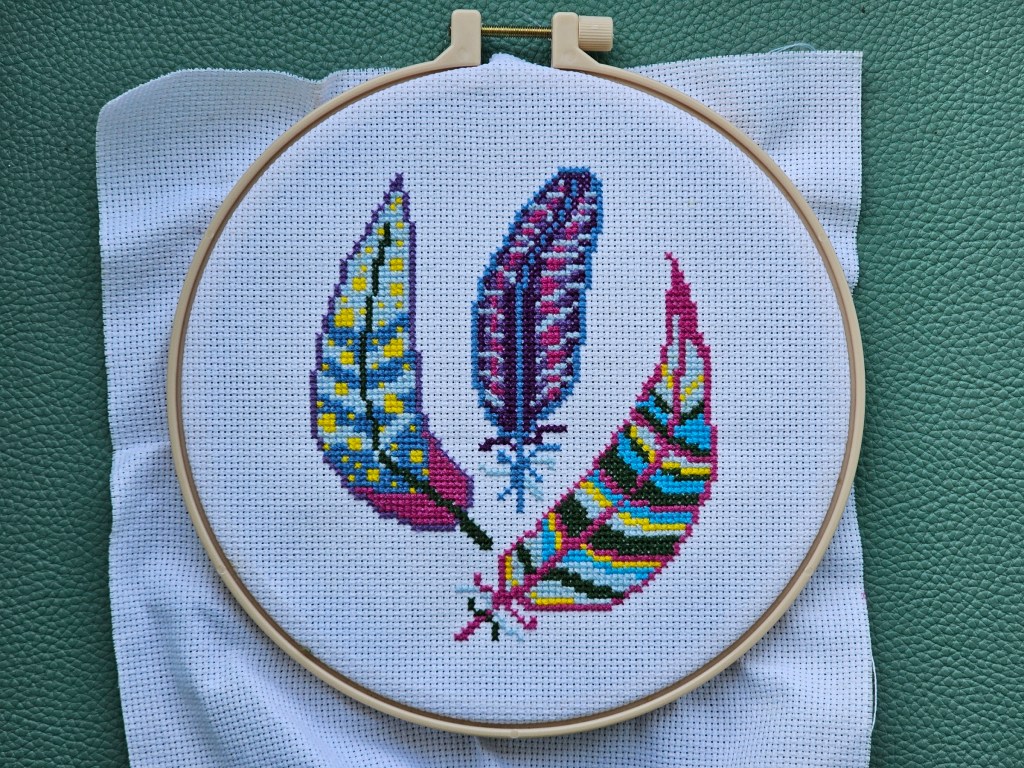 Finished cross stitch project featuring three pink, blue and purple feathers with yellow highlights.