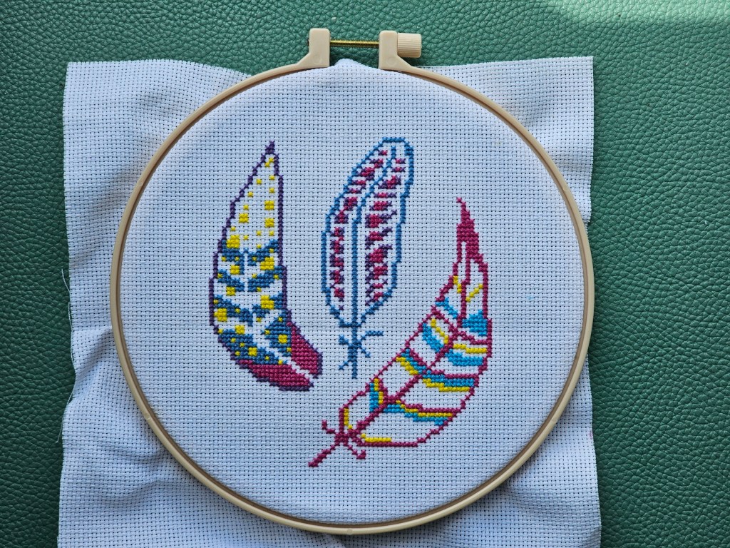 Partially finished cross stitch project featuring three pink, blue and purple feathers.