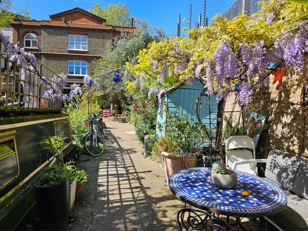A small garden with a table, a shed, and wisteria, located next to a canal path