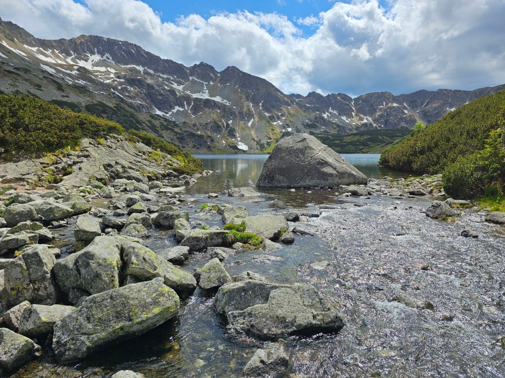 A stream leading to a mountain lake with some huge rocks in the water. Tall mountains can be seen in the background.