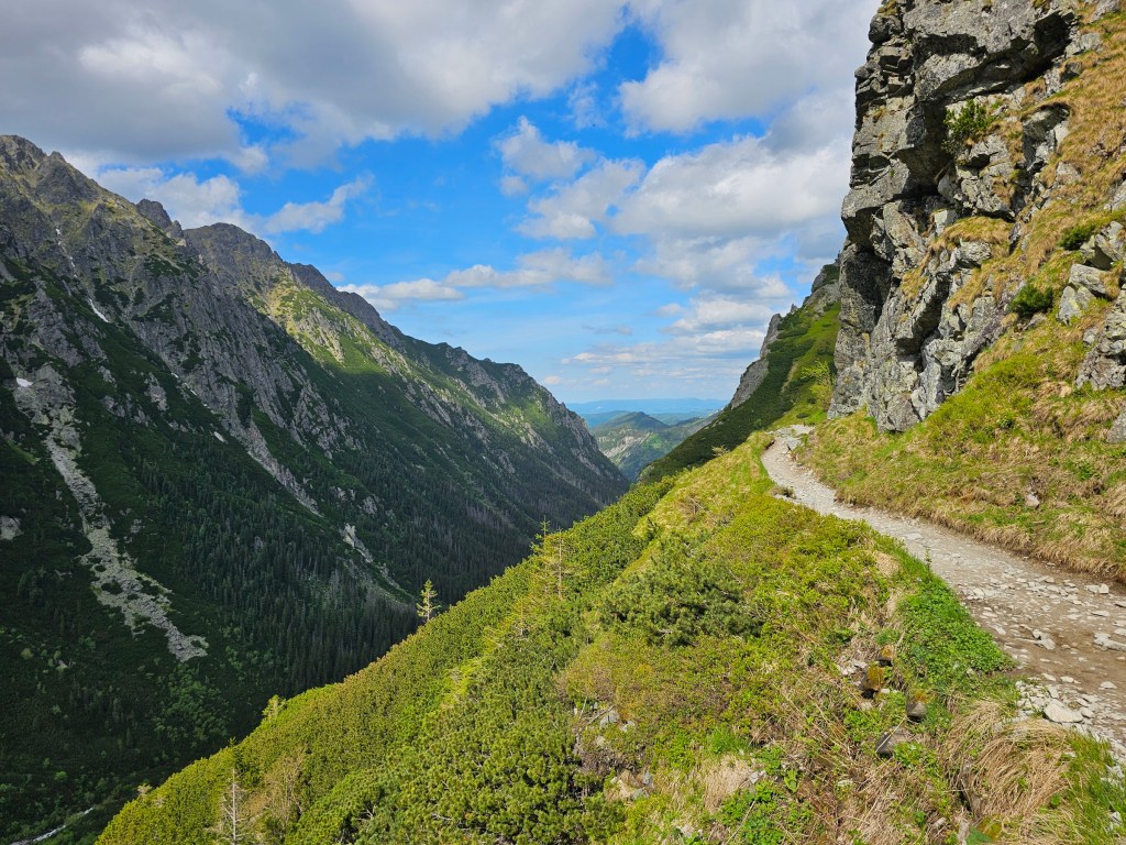 A narrow, gravel path on the side of a mountain. Other mountains can be seen nearby.