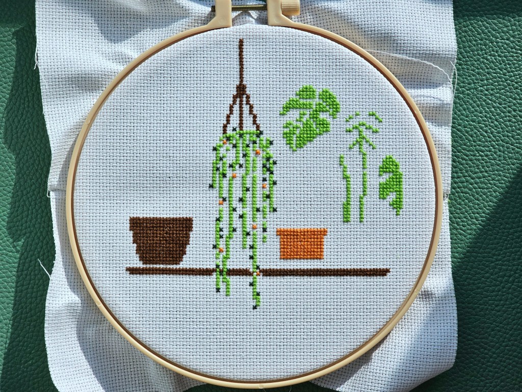 Partially finished cross stitch project featuring four house plants on a shelf