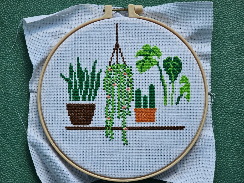 Nearly finished cross stitch project featuring four house plants on a shelf