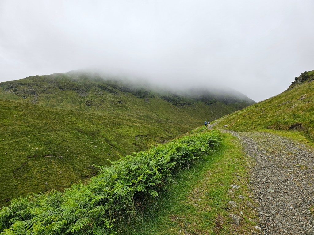 Green hills in a valley between Glenridding and Helvellyn. A mountain on the horizon is hiding in a thick cloud.