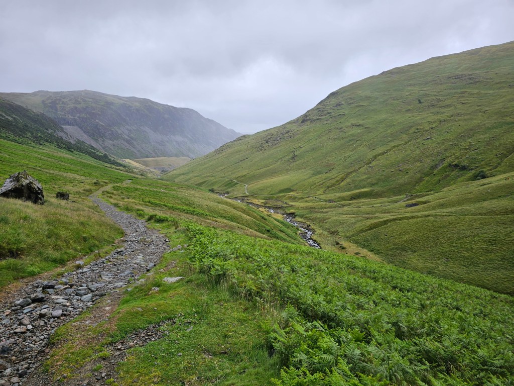 Green hills in a valley between Glenridding and Helvellyn with a small river at the bottom of the valley. Dense cloud covers the tops of the mountains.
