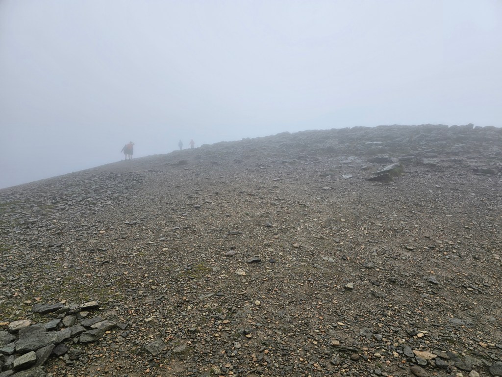The summit of Helvellyn, Lake District. There is dense fog and poor visibility. A few people can be seen in the distance.