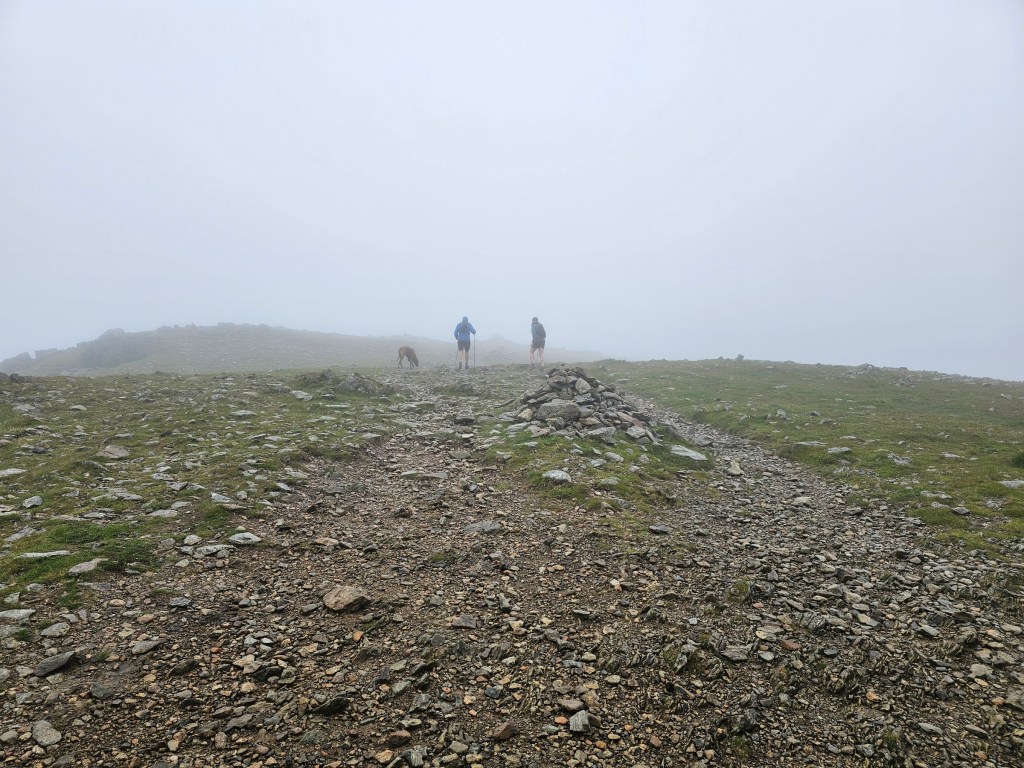 A wide gravel path near the summit of Helvellyn. Visibility is really poor. A couple of people and a dog can be seen in the distance.