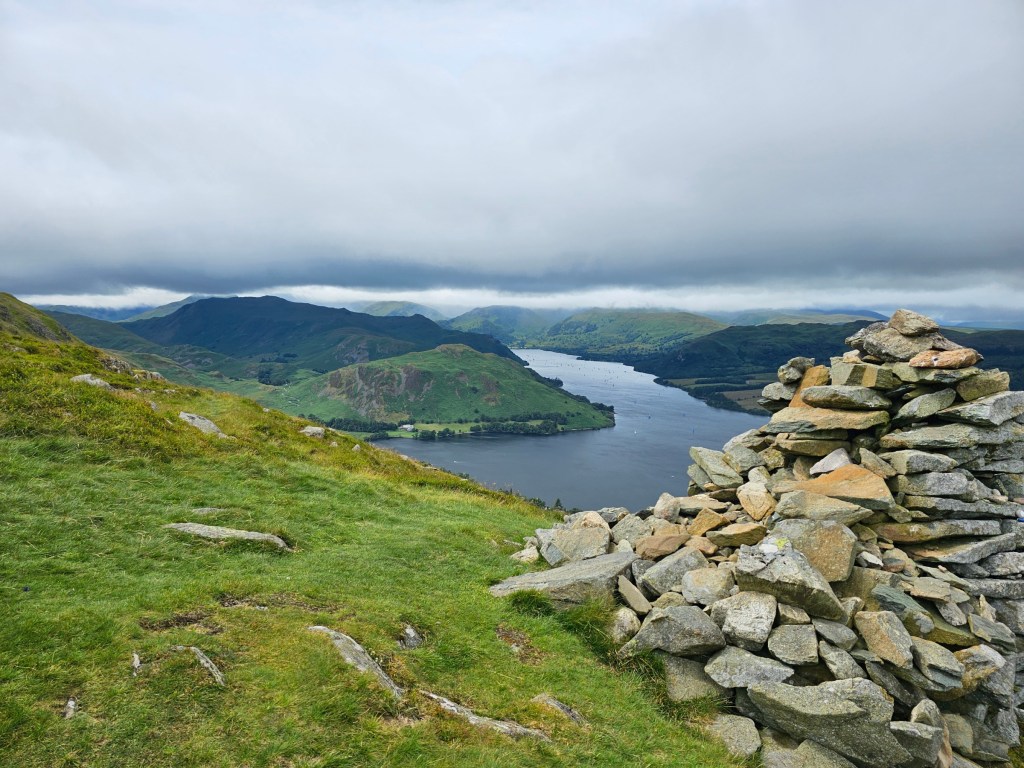 A pile of stones on top of a hill. A lake and other hills can be seen in the background