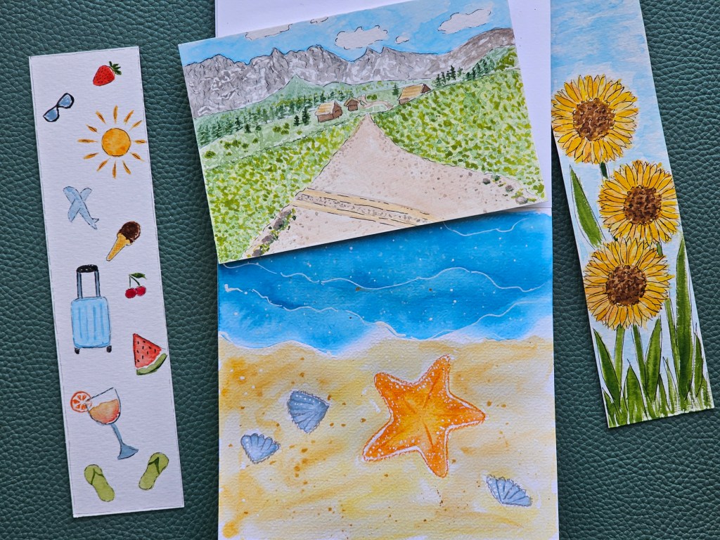 Handmade watercolour paintings in a summer theme. One postcard with a mountain landscape, a bookmark with sunflowers, a bookmark with summer icons, and a painting of a starfish on the beach