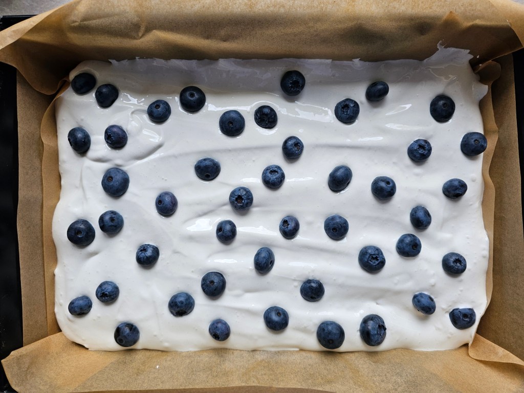 A tray with baked shortbread cake base, a layer of meringue, and blueberries on top - before baking