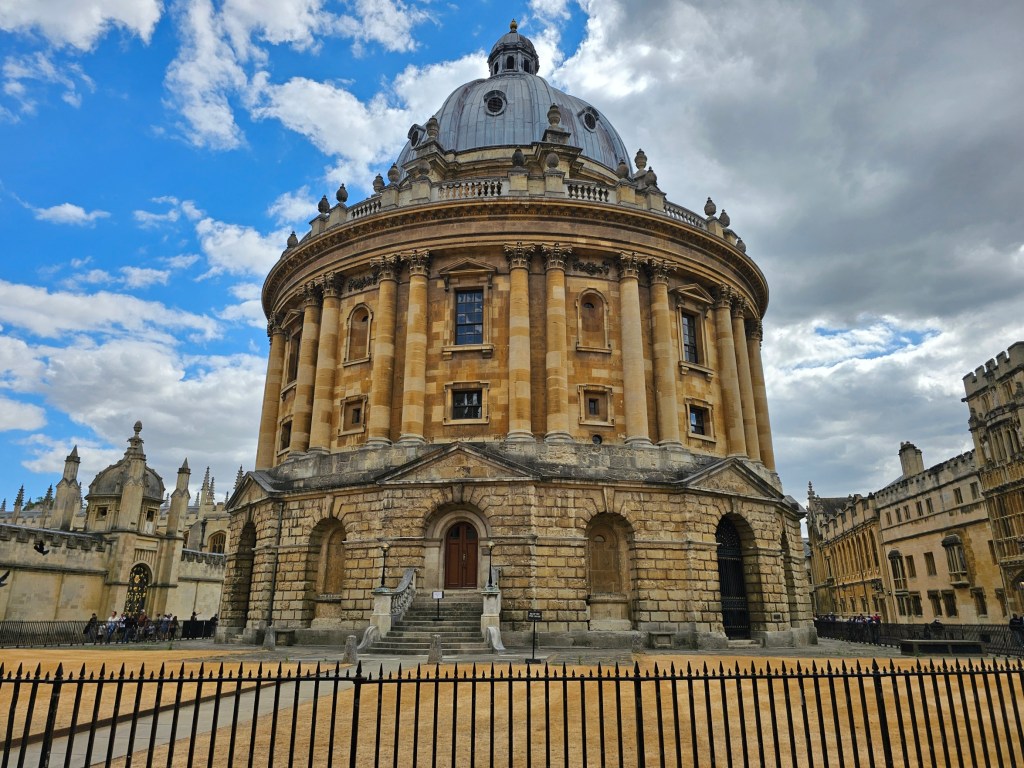 Bodleian Library, Oxford. The sky is largely blue, with some heavy clouds on the right side of the photo. The grass around the round building has dried up and turned to hay
