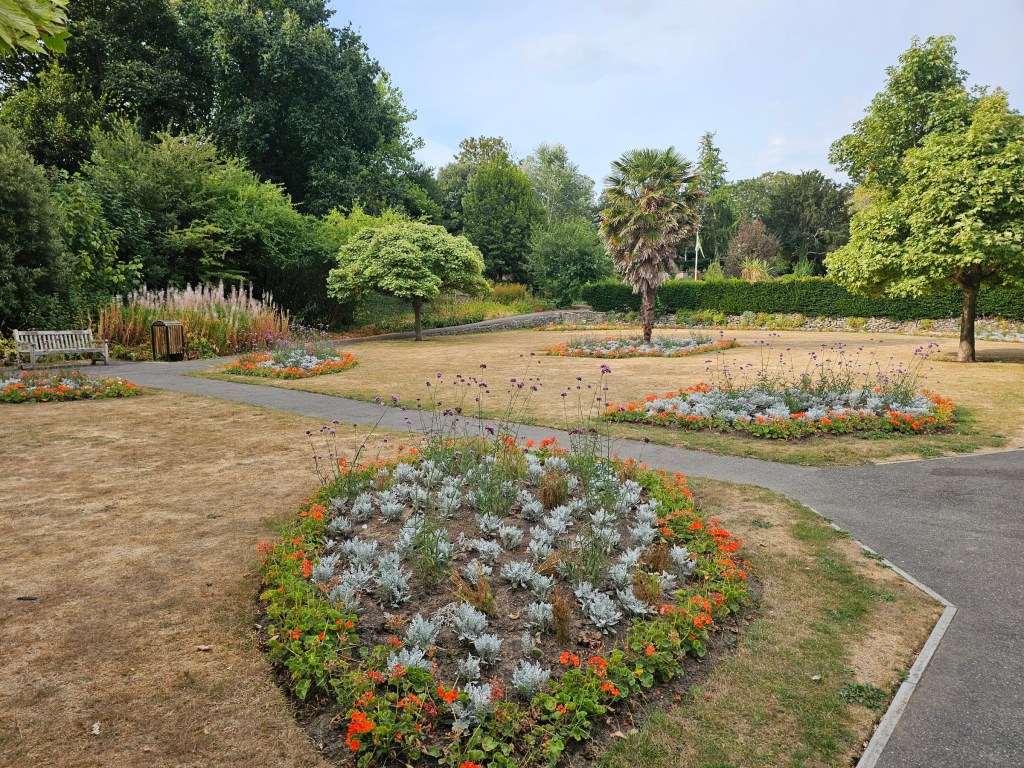 Kingsnorth Gardens in Folkestone. Flower beds have some colourful flowers, but the grass is dry and yellow.