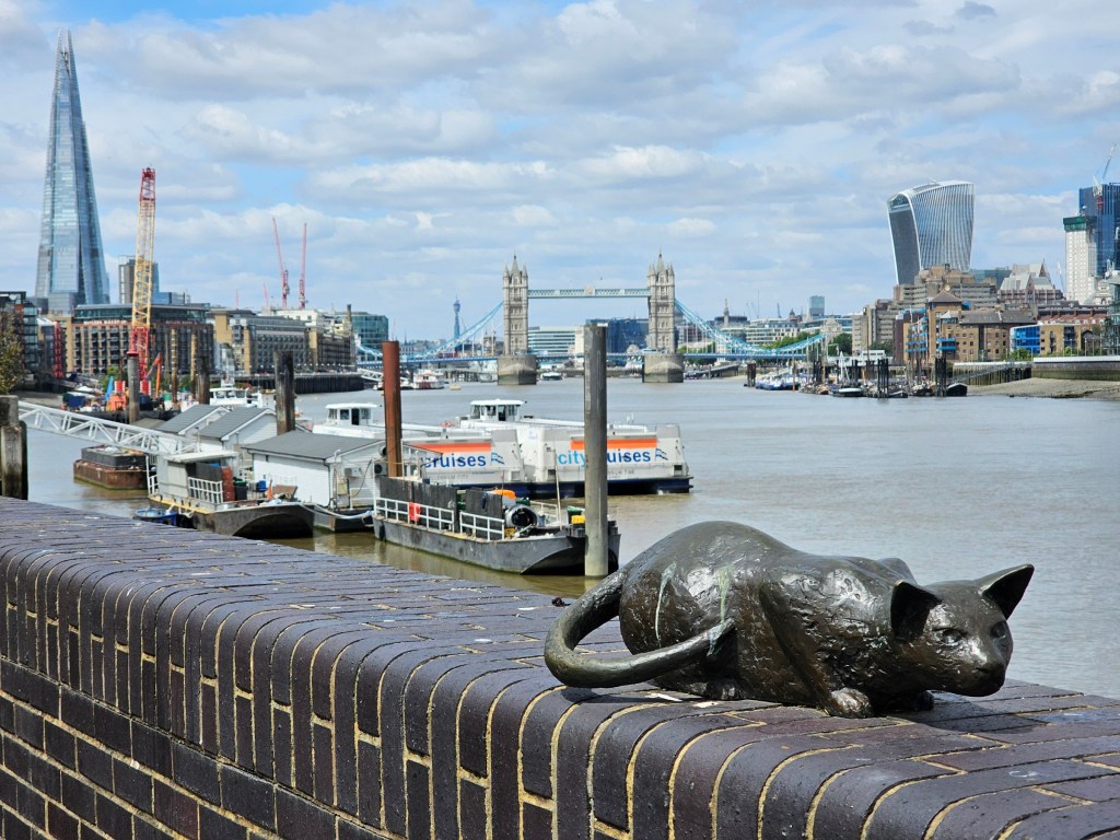 A view of Tower Bridge, the Shard, St Paul's Cathedral and the Walkie-Talkie building in London. In the foreground, there's a metal statue of a cat sitting on a wall.