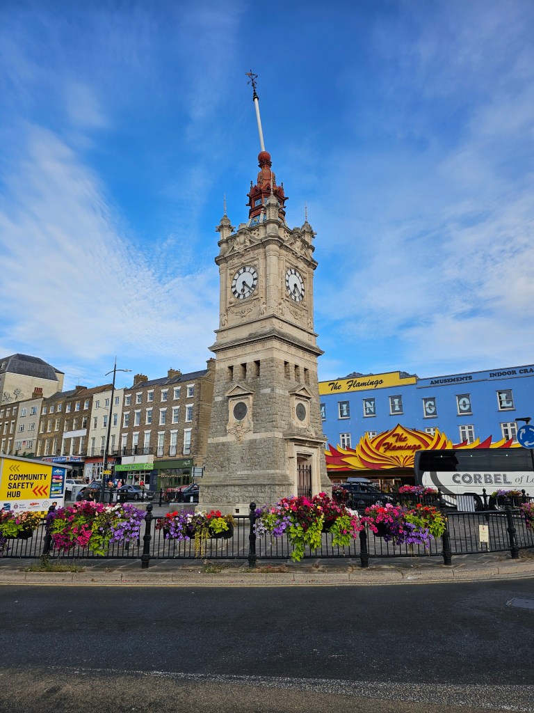 The clock tower in Margate, England. There's a blue building behind it, and colourful flowers on the railings around the tower