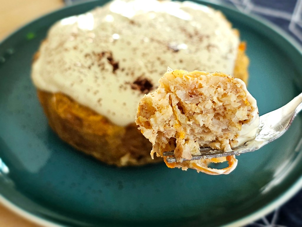 Baked carrot cake oats for one, on a plate, topped with yoghurt, and sprinkled with cocoa powder. A bite of the cake is held on a pastry fork.