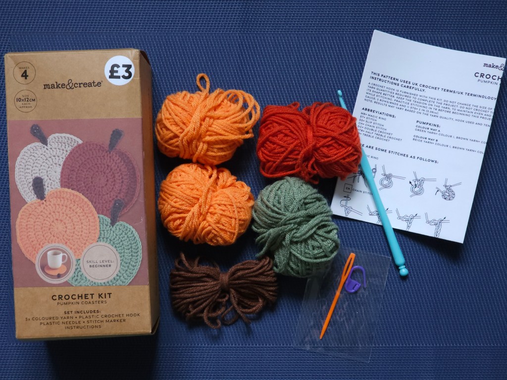 Contents of a crochet set for making crocheted pumpkin coasters. It includes five balls of yarn, a crochet hook, a plastic needle and a stitch marker.