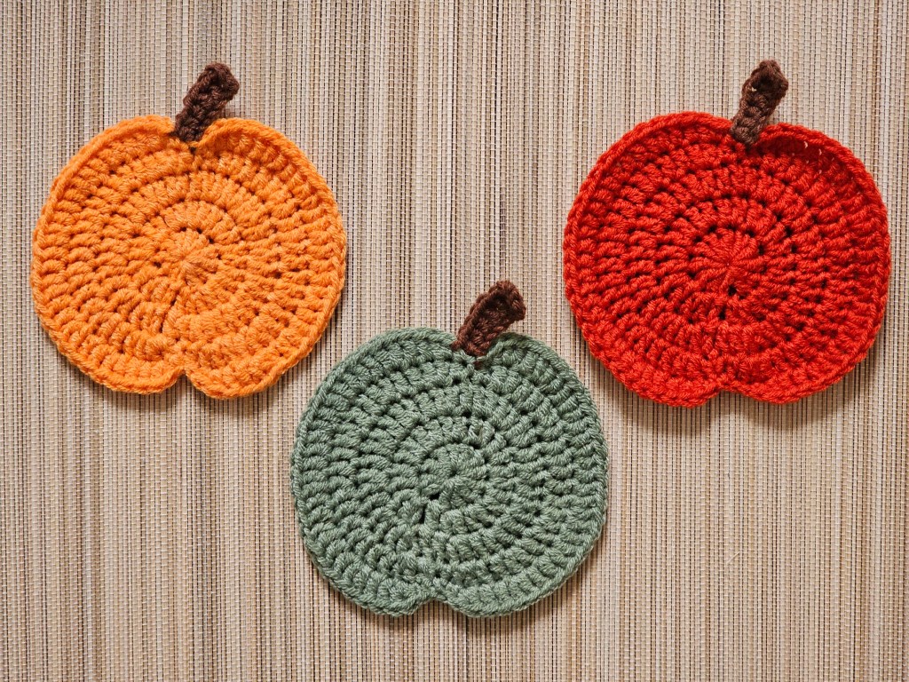 Three completed crocheted pumpkin coasters in orange, sage green, and red.