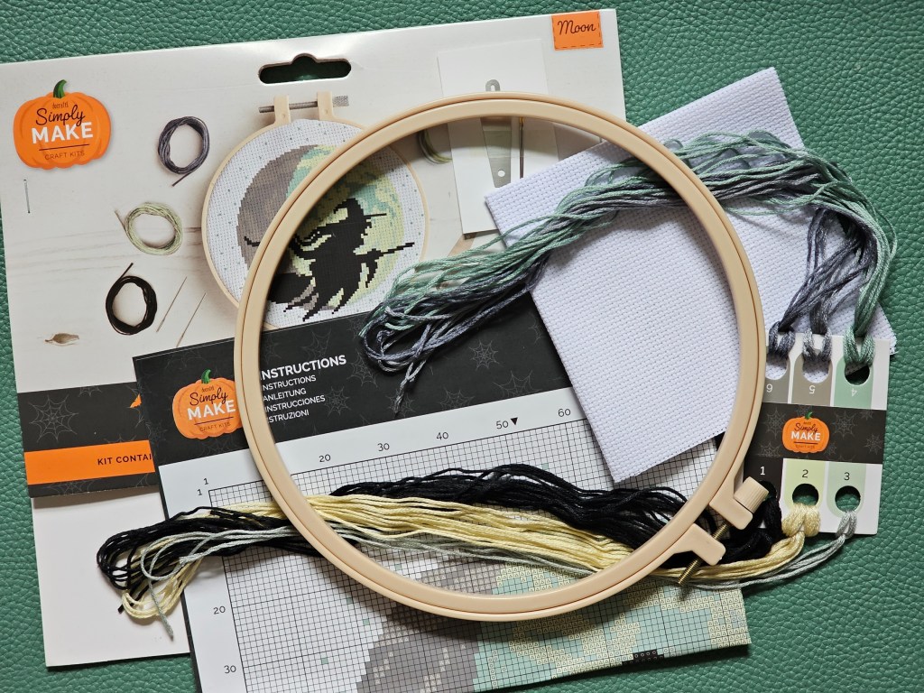 Contents of a DIY cross stitch set. It includes a large plastic embroidery hoop, a blank canvas, a needle, a threader, and threads in assorted colours.