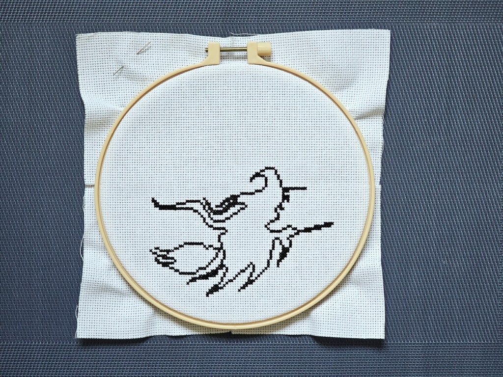Nearly finished cross stitch project depicting a witch on a flying broom in front of a large moon. Only a silhouette of the witch is sewn.