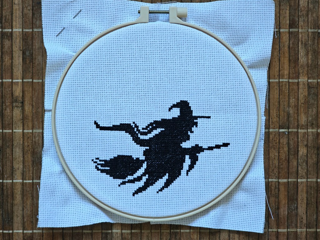 Nearly finished cross stitch project depicting a witch on a flying broom in front of a large moon. The witch is finished, the moon hasn't been started.