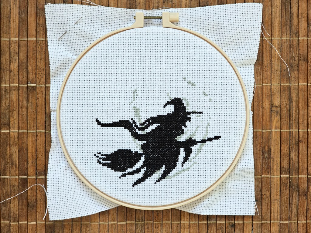 Nearly finished cross stitch project depicting a witch on a flying broom in front of a large moon. The witch is finished, the moon is only started.