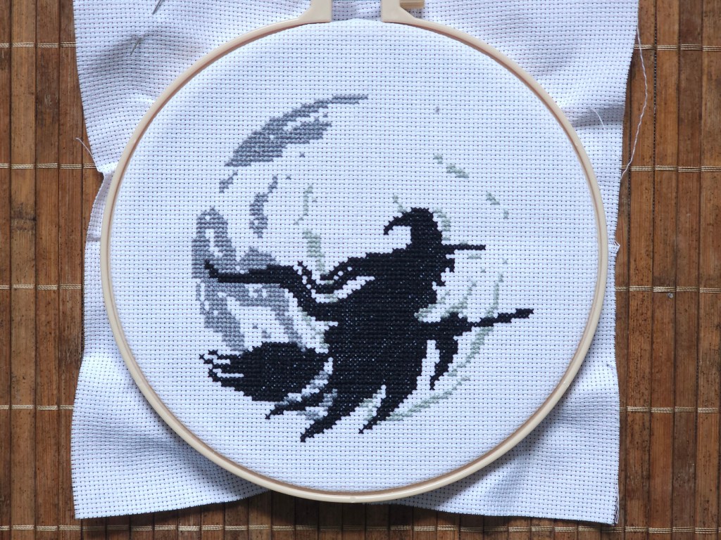 Nearly finished cross stitch project depicting a witch on a flying broom in front of a large moon. The witch is finished, the moon is only started.