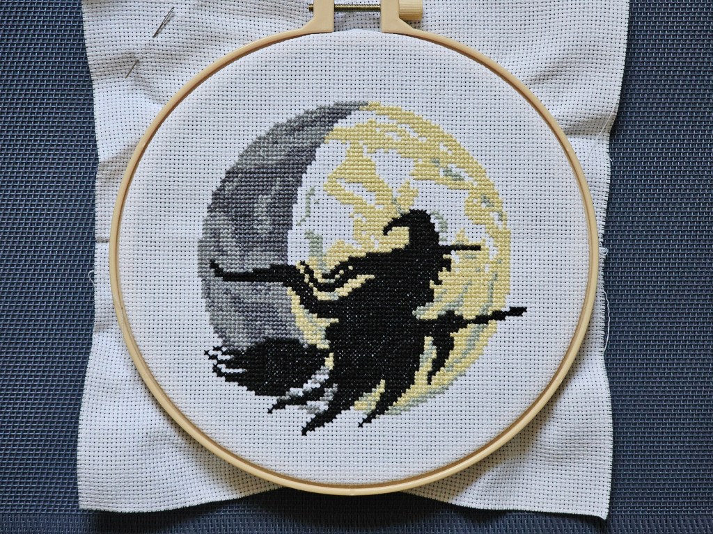 Nearly finished cross stitch project depicting a witch on a flying broom in front of a large moon