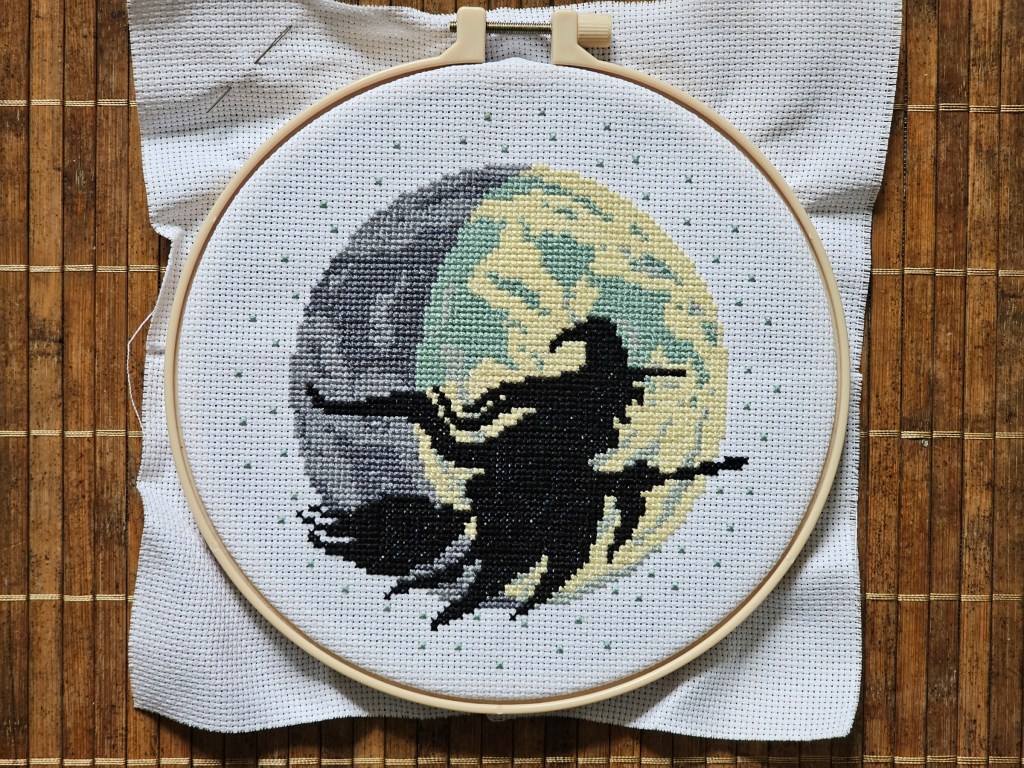 Finished cross stitch project depicting a witch on a flying broom in front of a large moon